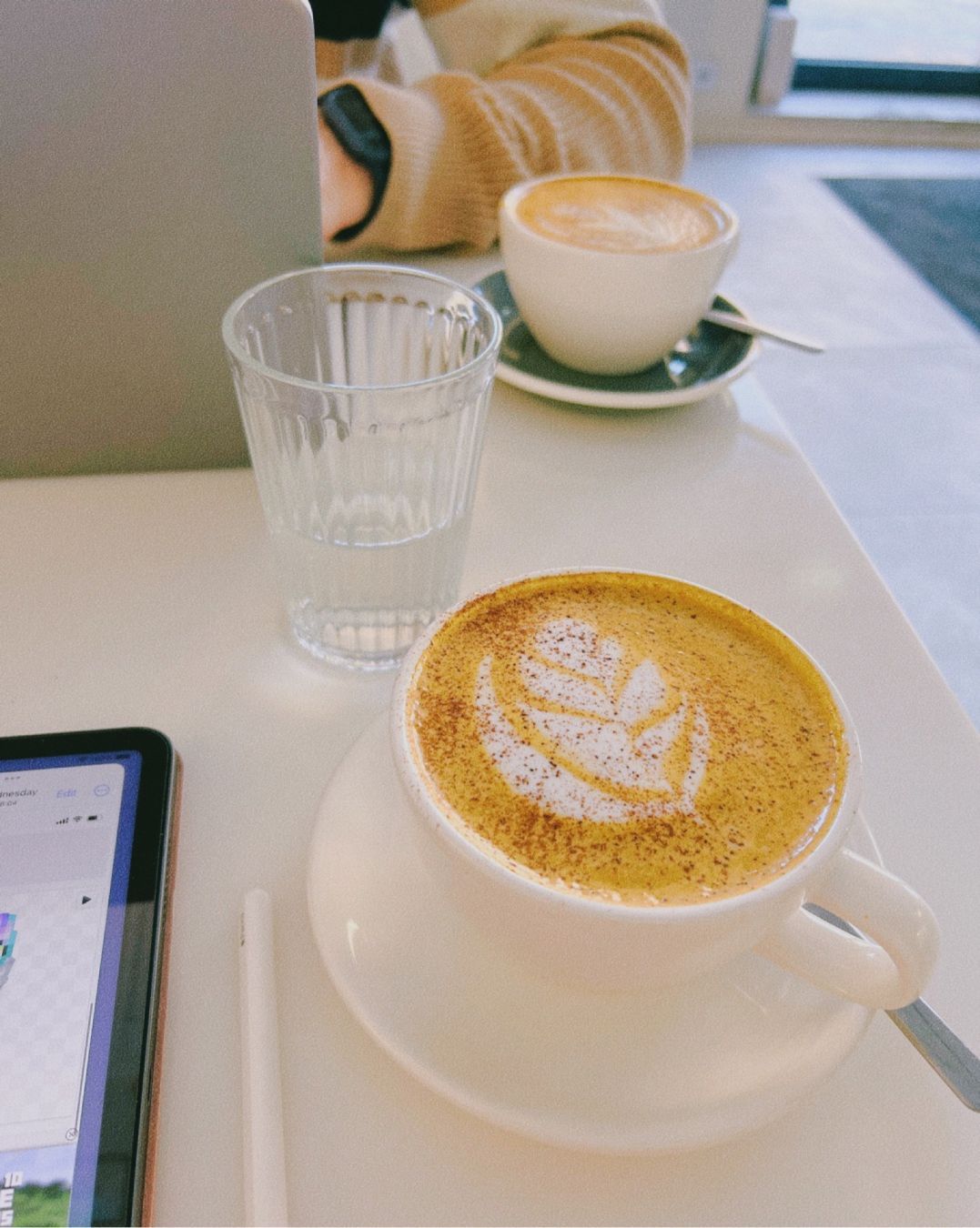 partially visible table at a café, with two pumpkin spice latte’s, a glass of water, a tablet, and a laptop